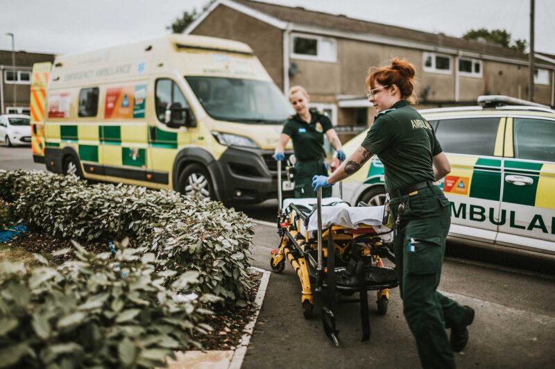 Ambulance crew pushing a stretcher into a house at a housing estate with an ambulance and RRV in the background