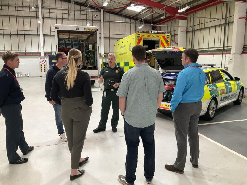 A paramedic in uniform speaks to a small group of people standing in front of a selection of emergency vehicles