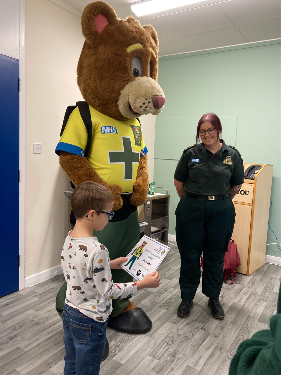 A young boy receives a certificate from an ambulance worker and bear mascot