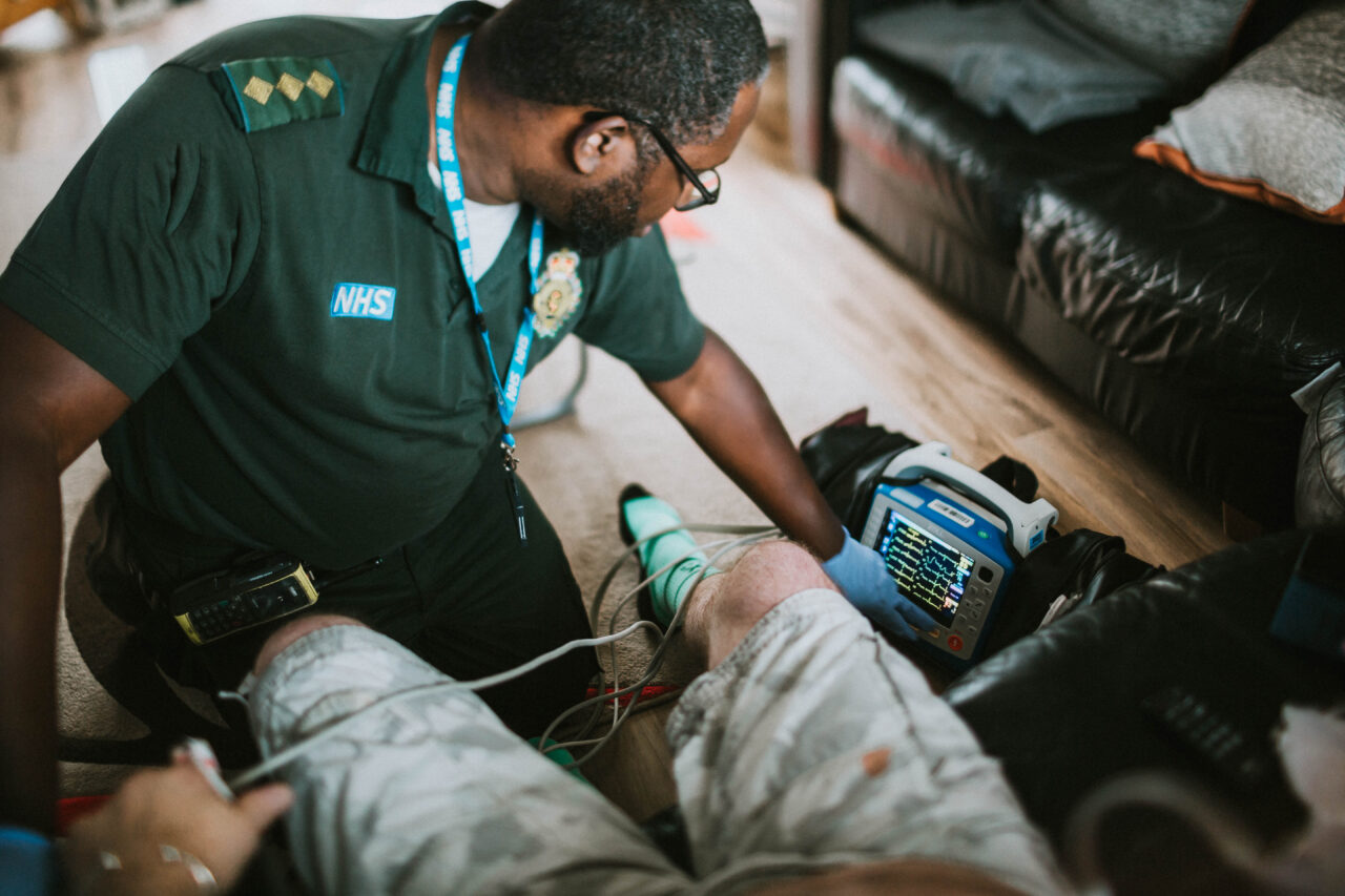 Paramedic checks the display on a monitor whilst assessing a patient in their home