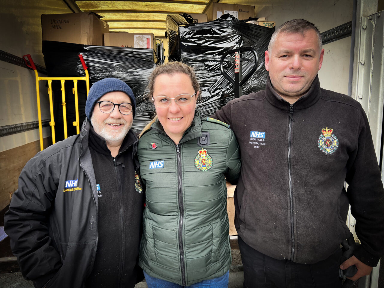 Three people standing arm-in-arm in front of a lorry load of supplies