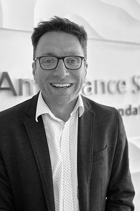 Black and white portrait photo of Craig Ellis in a suit and shirt standing in front of the corporate signage