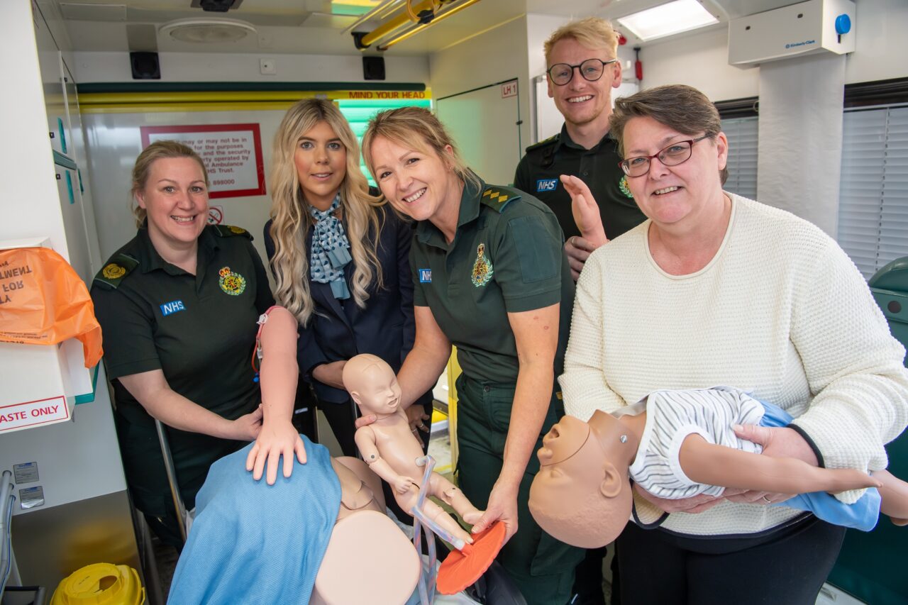 A group of five people, including three paramedics in uniform, holding training equipment inside an ambulance