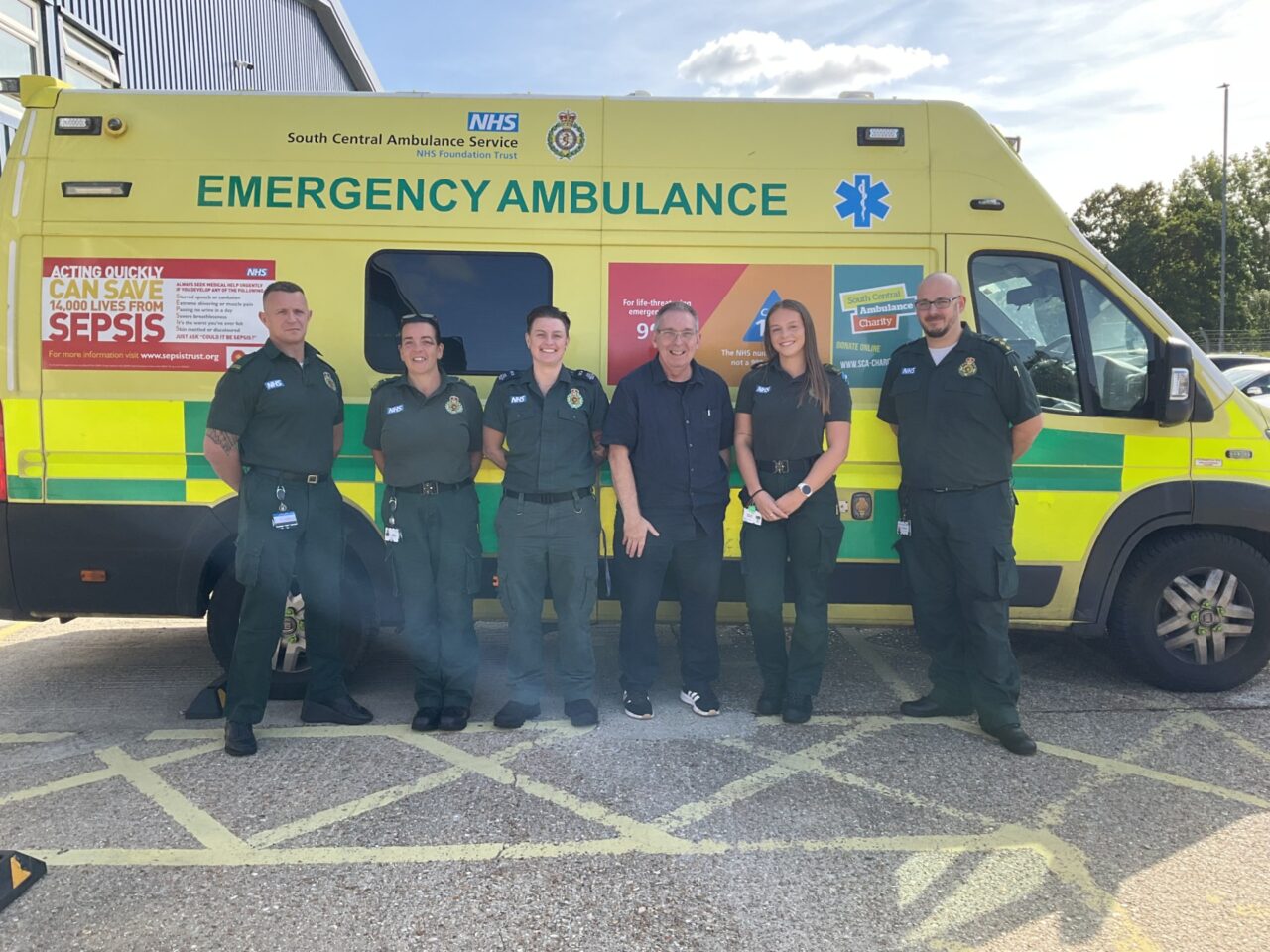 Five members of the ambulance service in uniform with a member of the public standing in front of an emergency ambulance