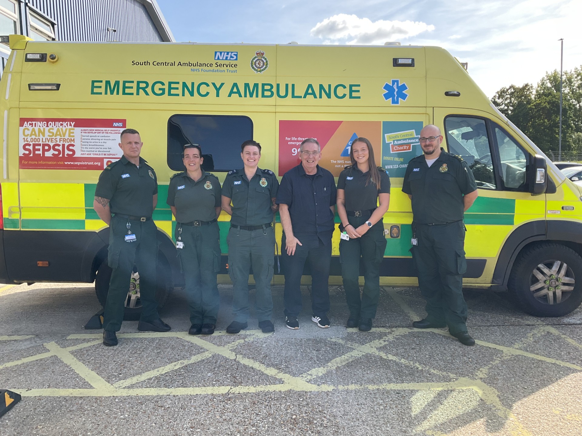 Five members of the ambulance service in uniform with a member of the public standing in front of an emergency ambulance