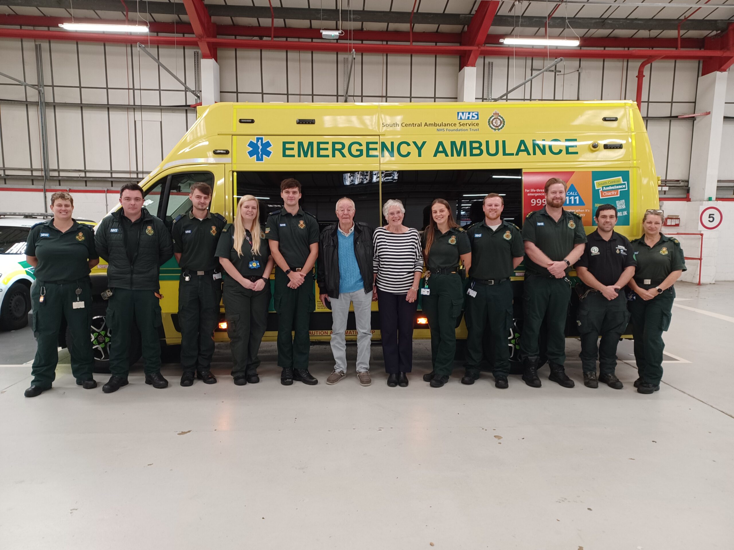A line of ambulance and staff volunteers in green uniforms standing in front of an ambulance with a male and female member of the public in the middle of the twelve strong group.