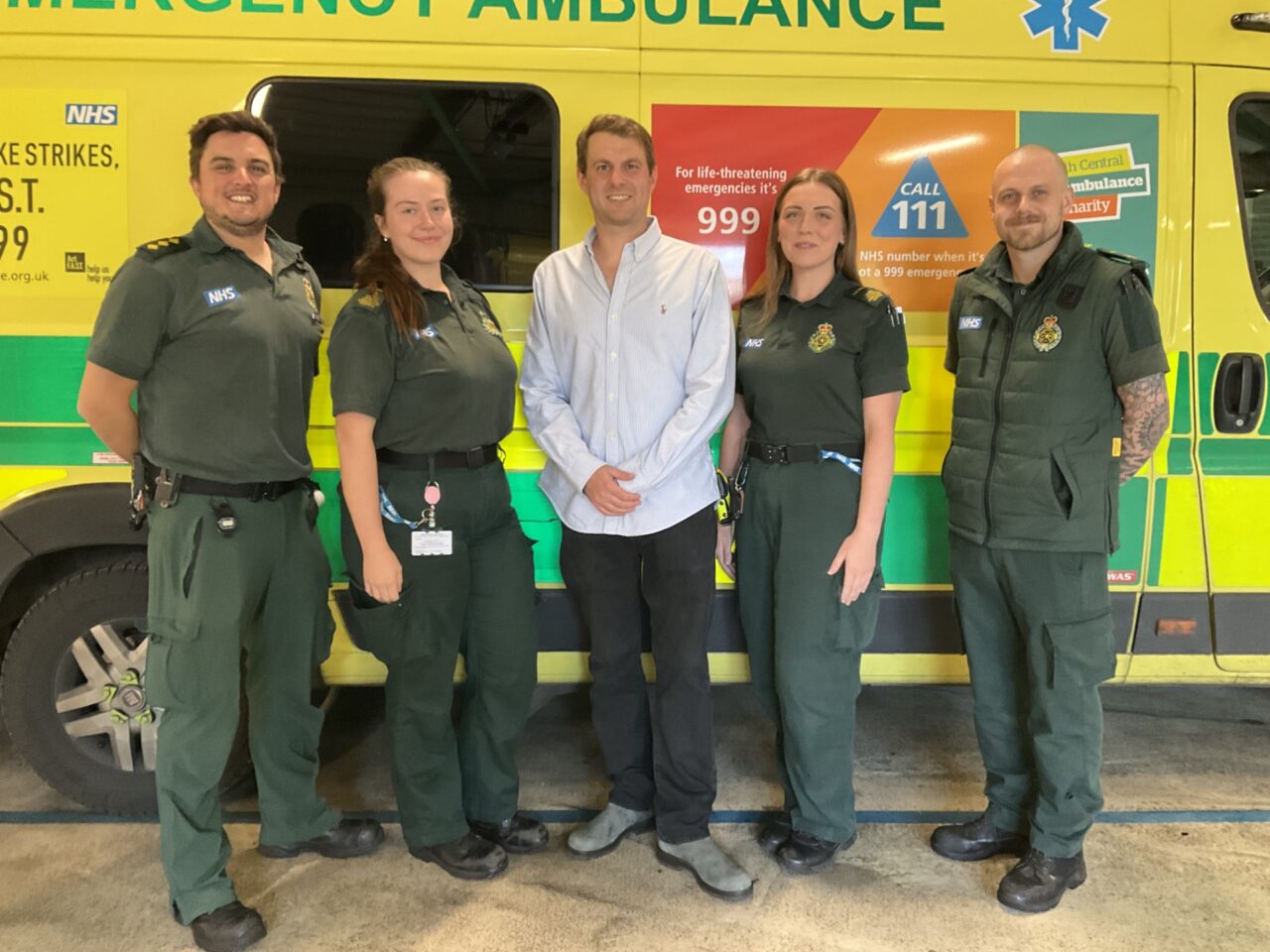 Male member of the public standing in the middle of a group of five people in front of an ambulance with a male and female member of staff in green ambulance uniforms either side of him