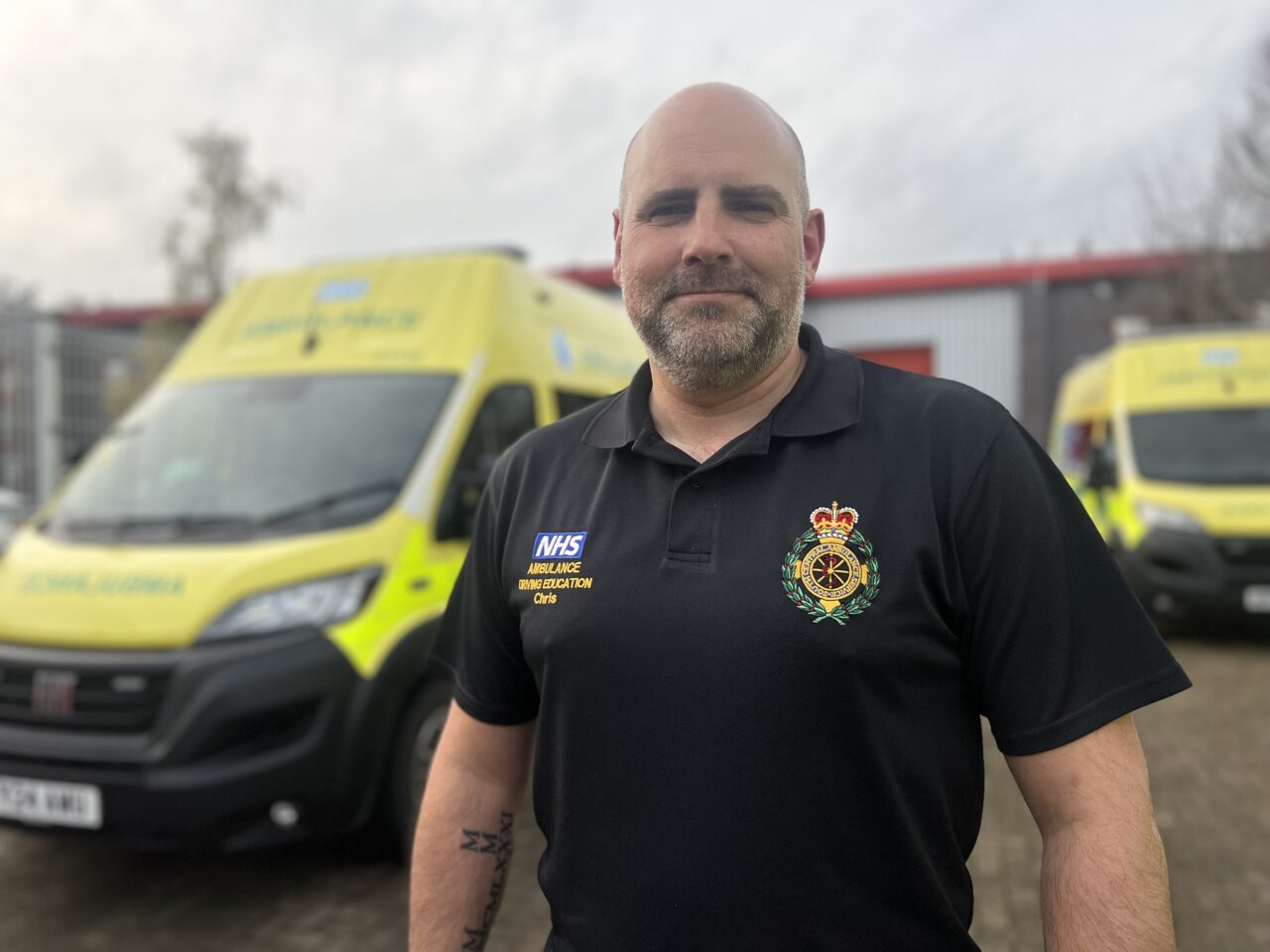 An ambulance driver training instructor standing outside with emergency ambulance vehicles blurred in the background