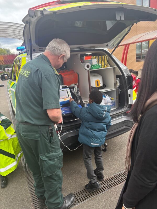 A male paramedic in green uniform standing at the back of an emergency response car which has its boot open and he is showing a reception class pupil some of the ambulance kit contained in it.
