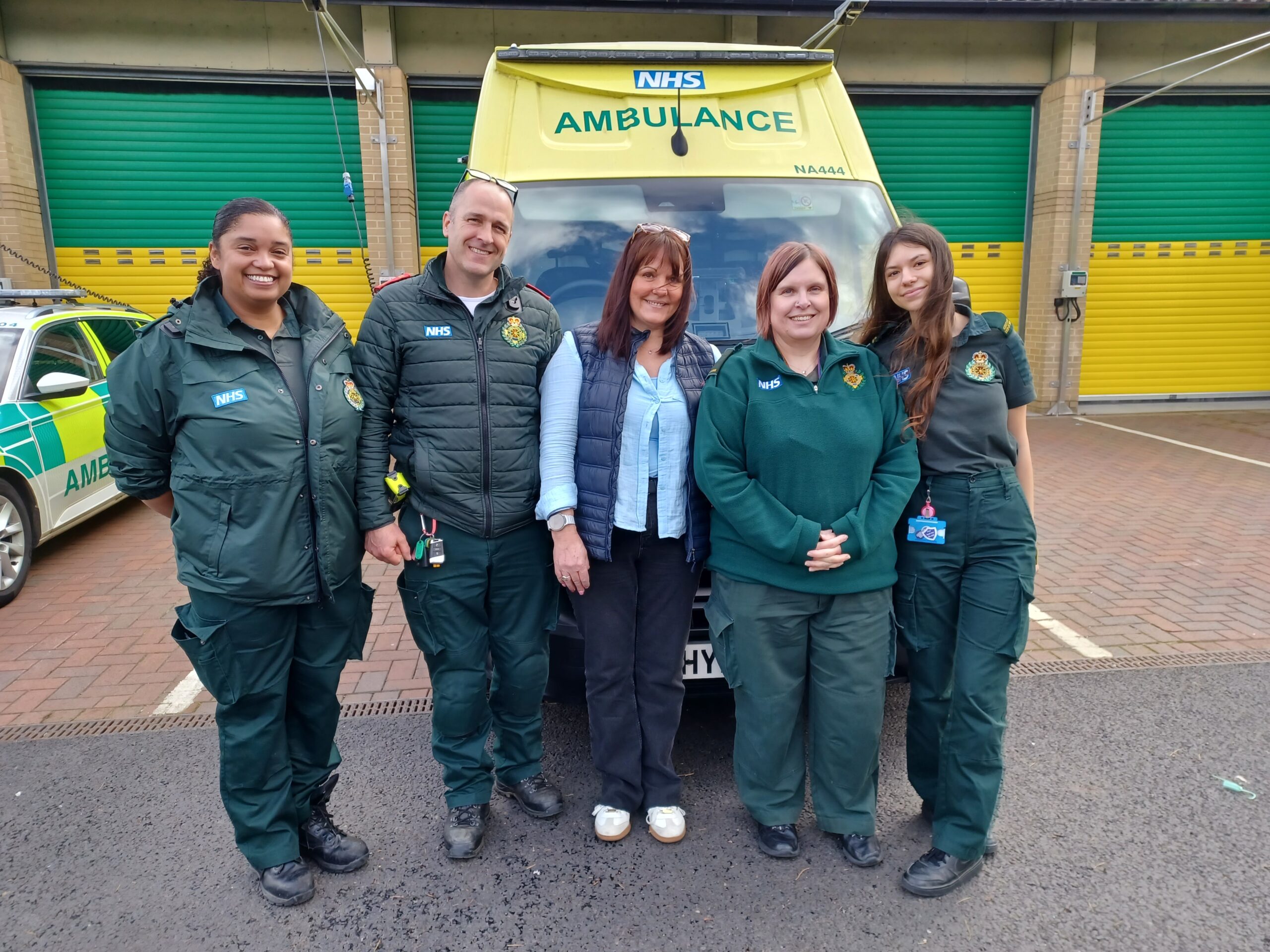 Four ambulance service staff in green uniform with an adult female standing in front of an ambulance