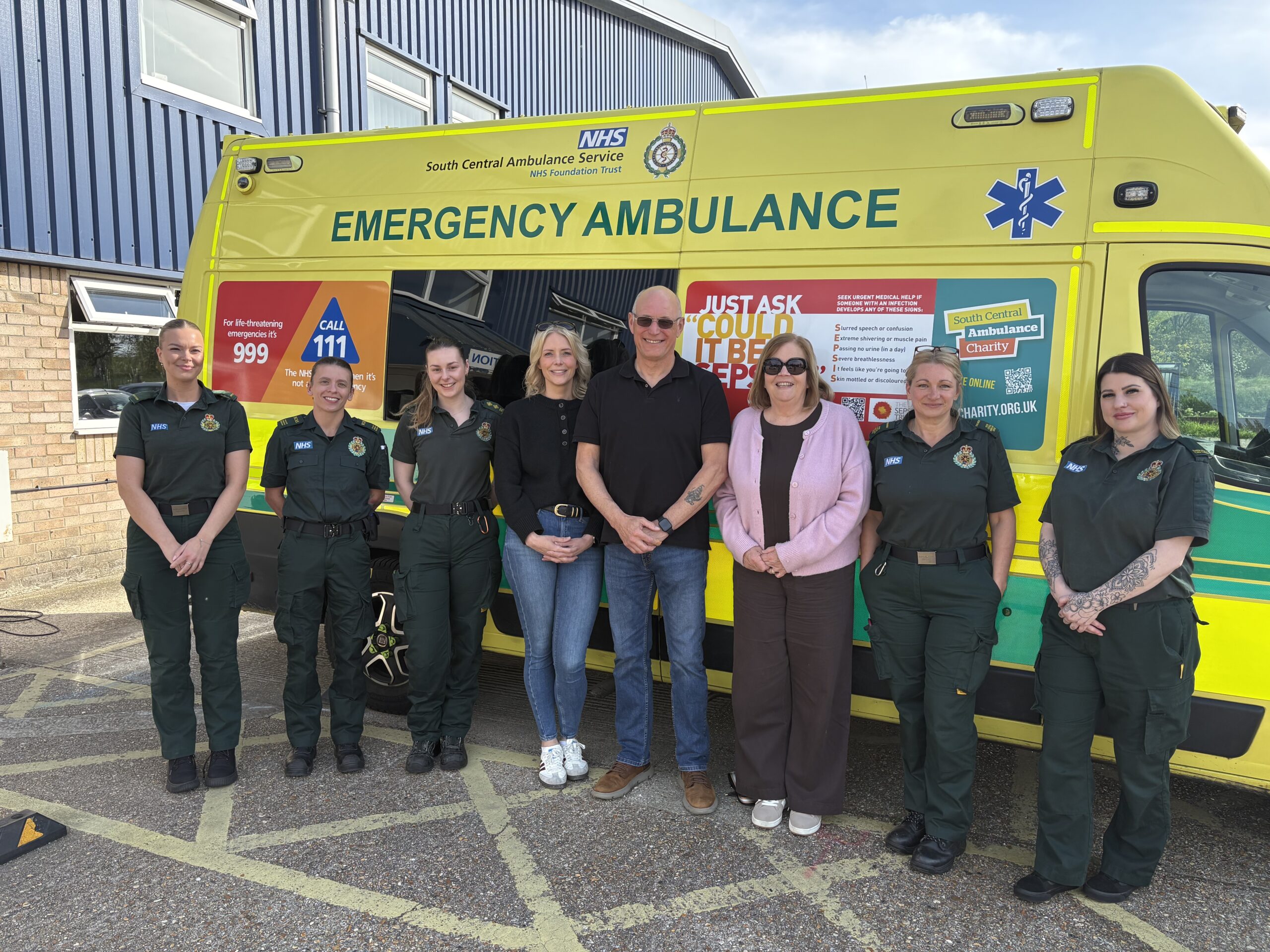 A man, woman and their adult daughter standing in front of an ambulance flanked by five female ambulance service staff in green uniform.