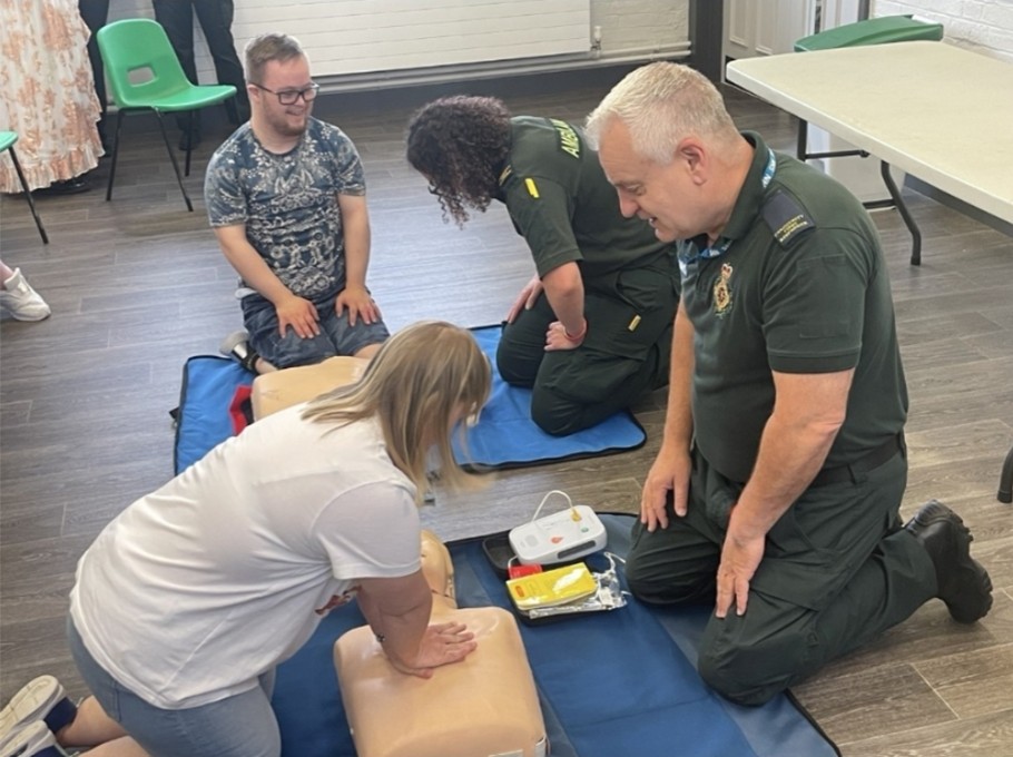 Two ambulance staff kneeling on the floor helping two members of the public learn how to carry out CPR using mannequins