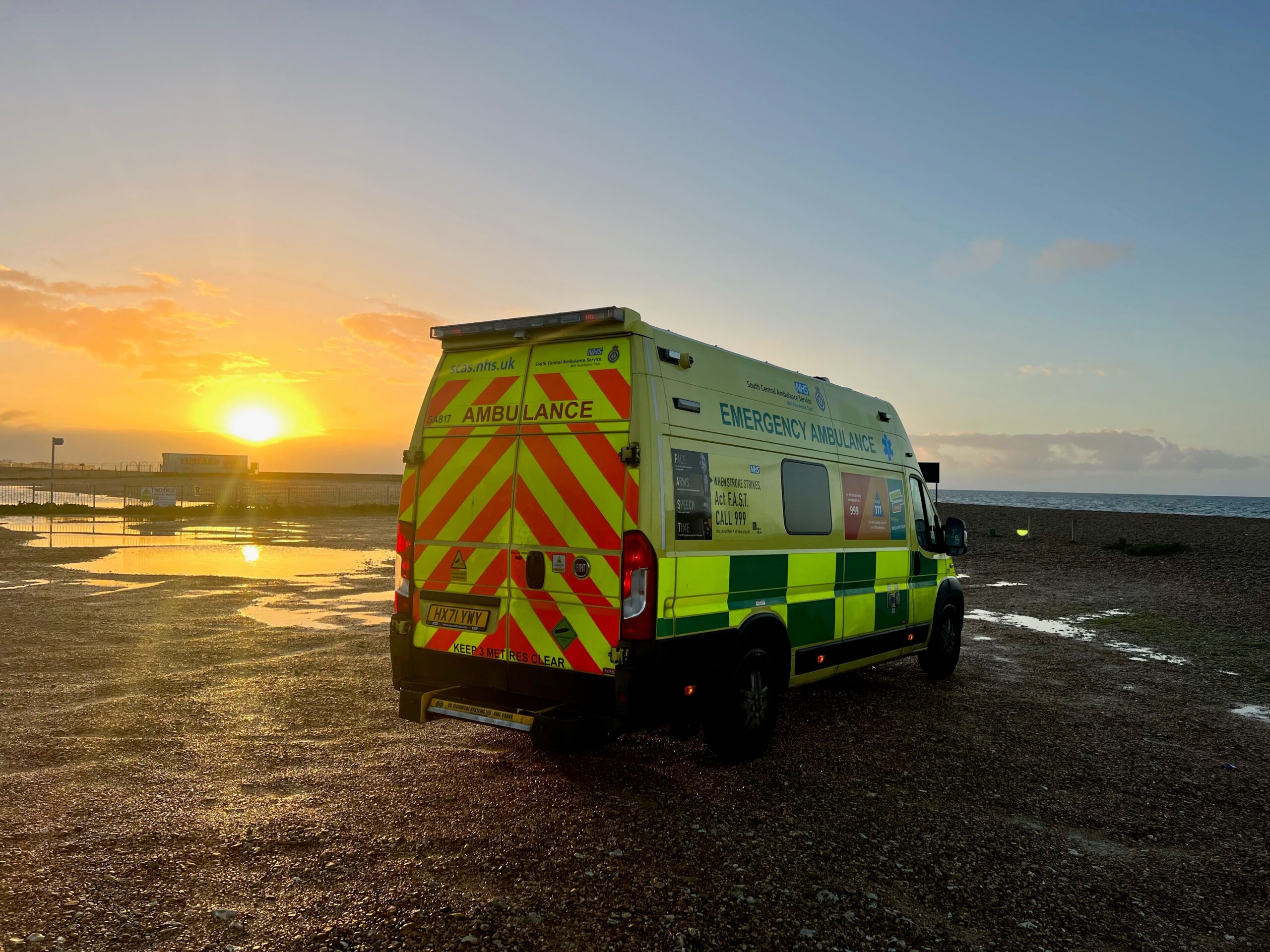 An ambulance parked on gravel by a small sea wall overlooking the coast as the sun rises on the horizon.