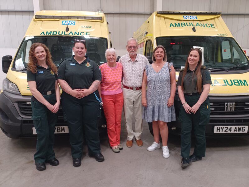Three ambulance staff in green uniform and three members of the public are standing in an ambulance garage in front of two ambulance vehicles
