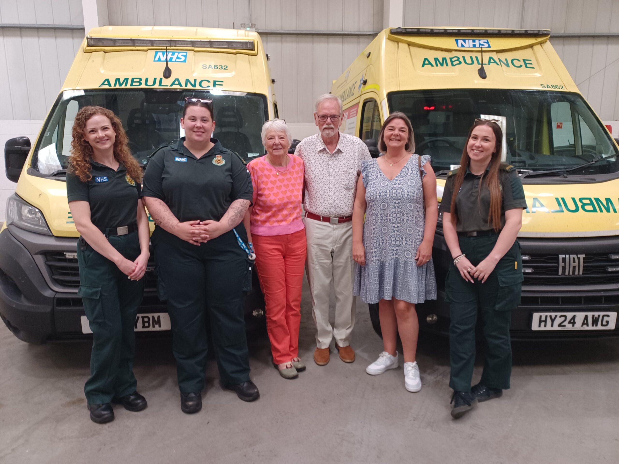 Three ambulance staff in green uniform and three members of the public are standing in an ambulance garage in front of two ambulance vehicles