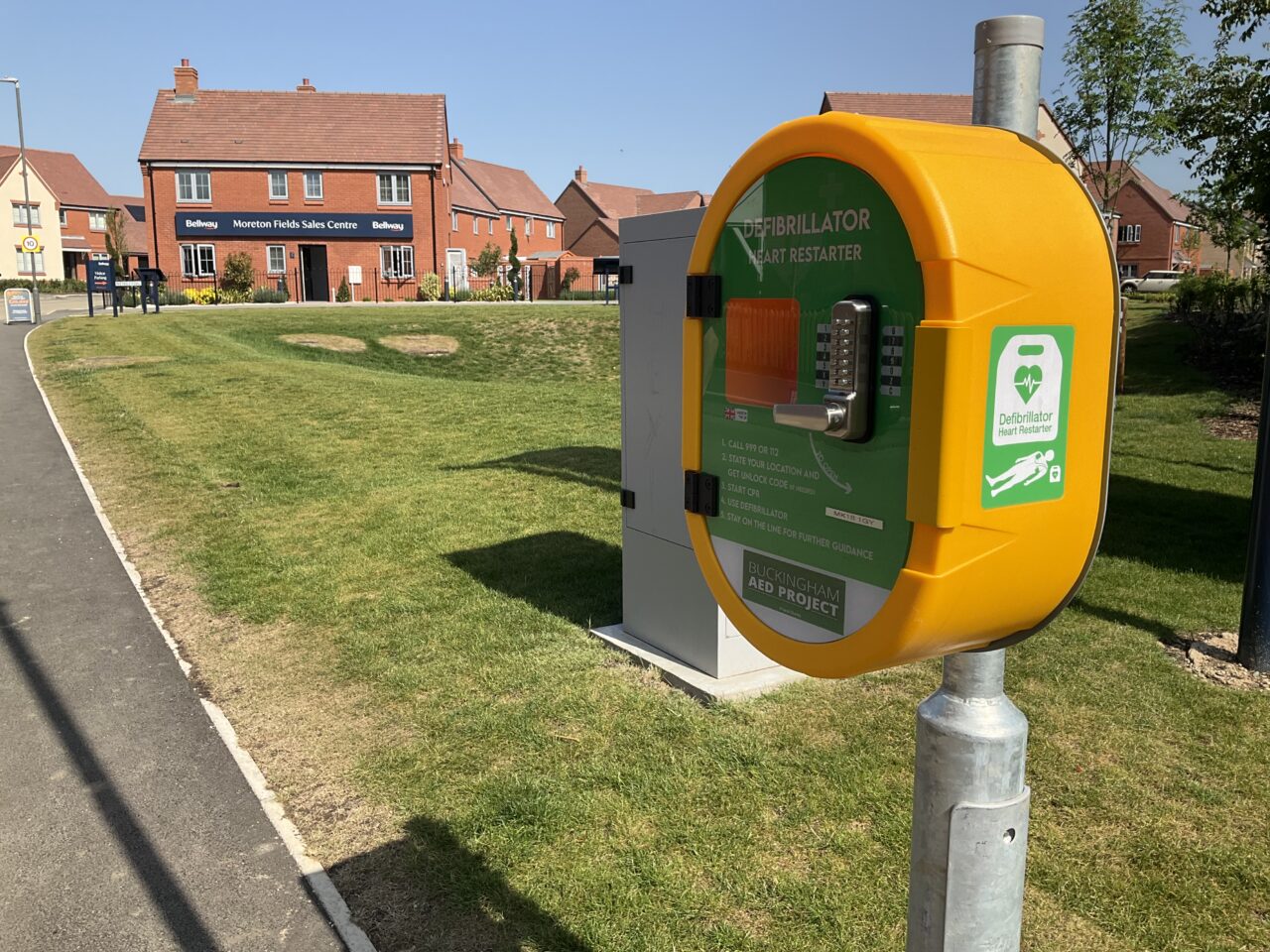 A yellow locked cabinet housing a defibrillator attached to a metal pole on a new housing estate