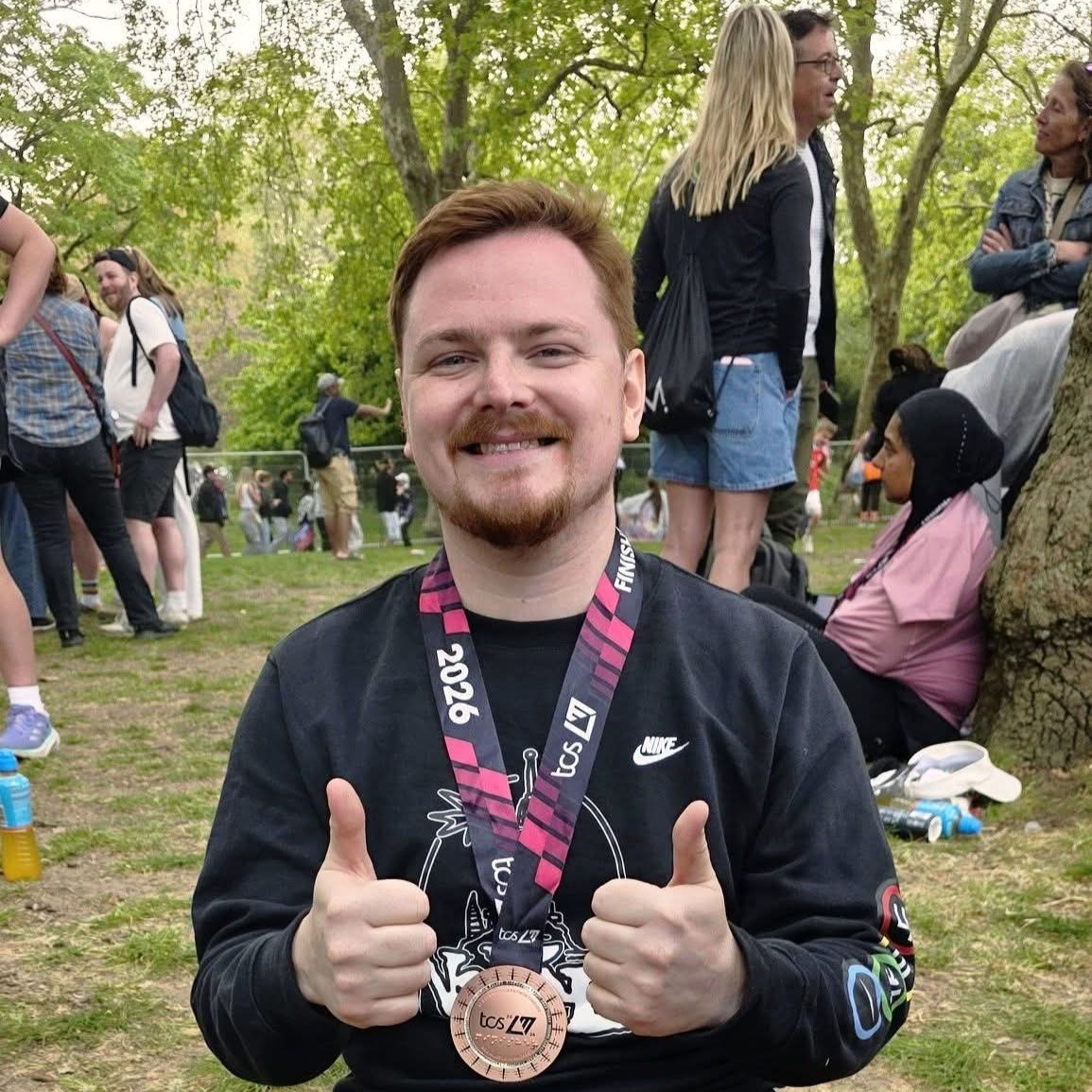 Luke Daskalakis 4 A man sitting on the grass, smiling with two thumbs up and wearing a London Marathon Finisher Medal