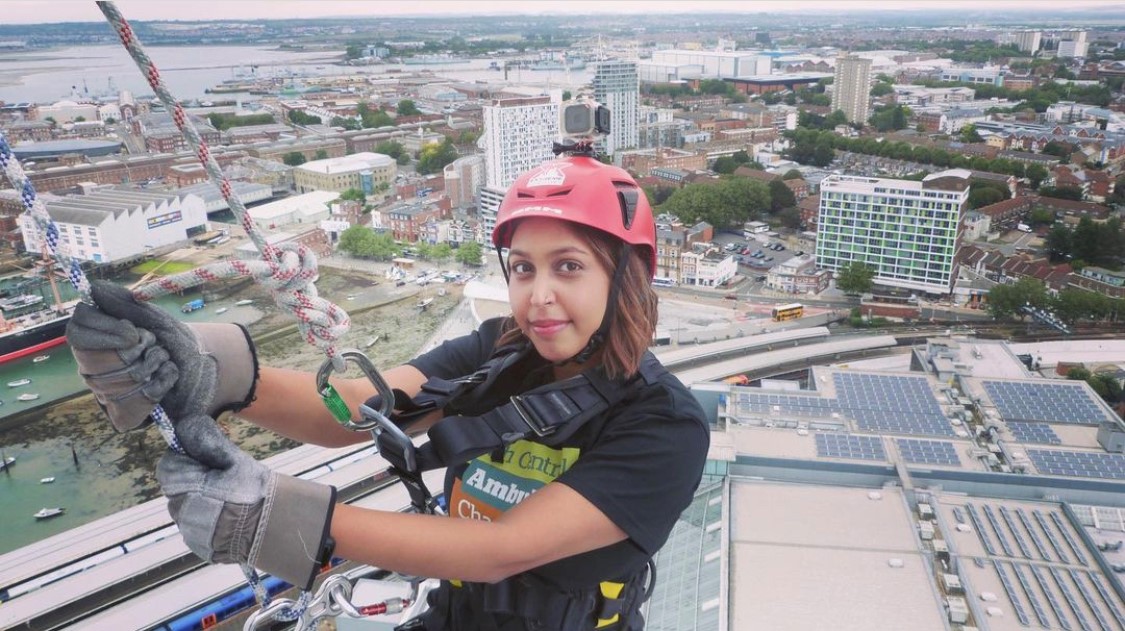 A woman in a safety harness, hard hat, wearing gloves and holding on to a rope. She is looking at the camera from the top of the 100m Spinnaker Tower in Portsmouth as she starts to abseil down