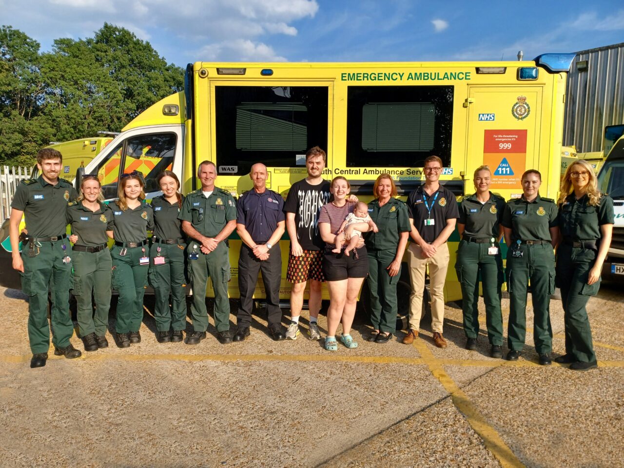A group of 10 ambulance service responders and one firefighter stand in a line in front of an ambulance. In the centre of the group is a young married couple with the female holding a small baby.
