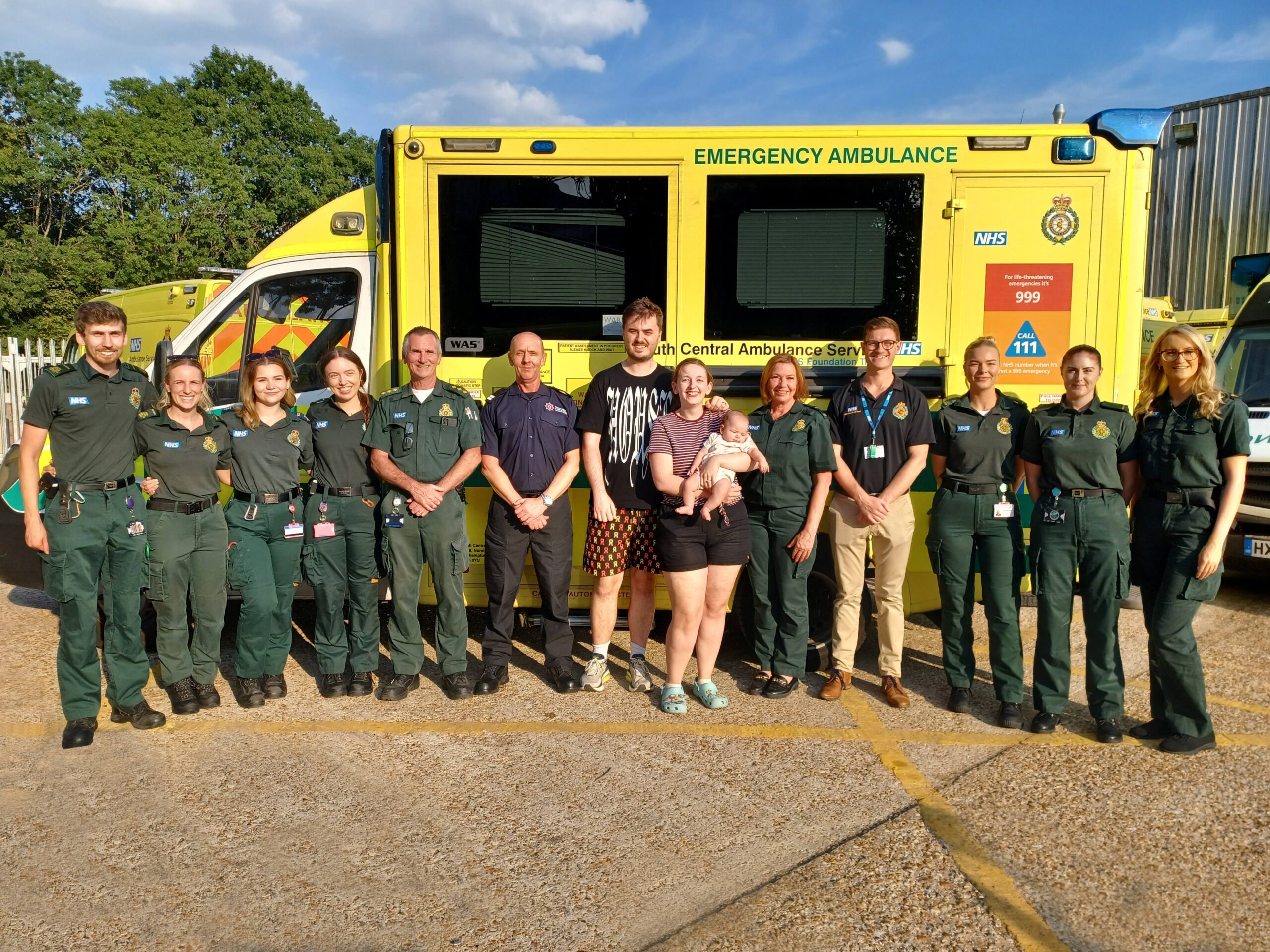 A group of 10 ambulance service responders and one firefighter stand in a line in front of an ambulance. In the centre of the group is a young married couple with the female holding a small baby.