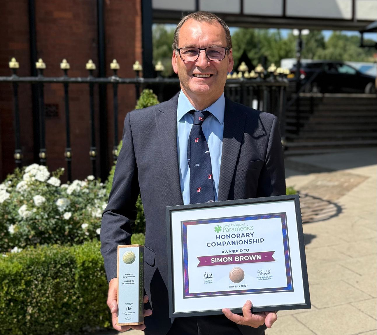 Simon Brown CoP Award July 2026 A smiling man in a suit is standing outside holding a framed certificate in one hand and an award in the other