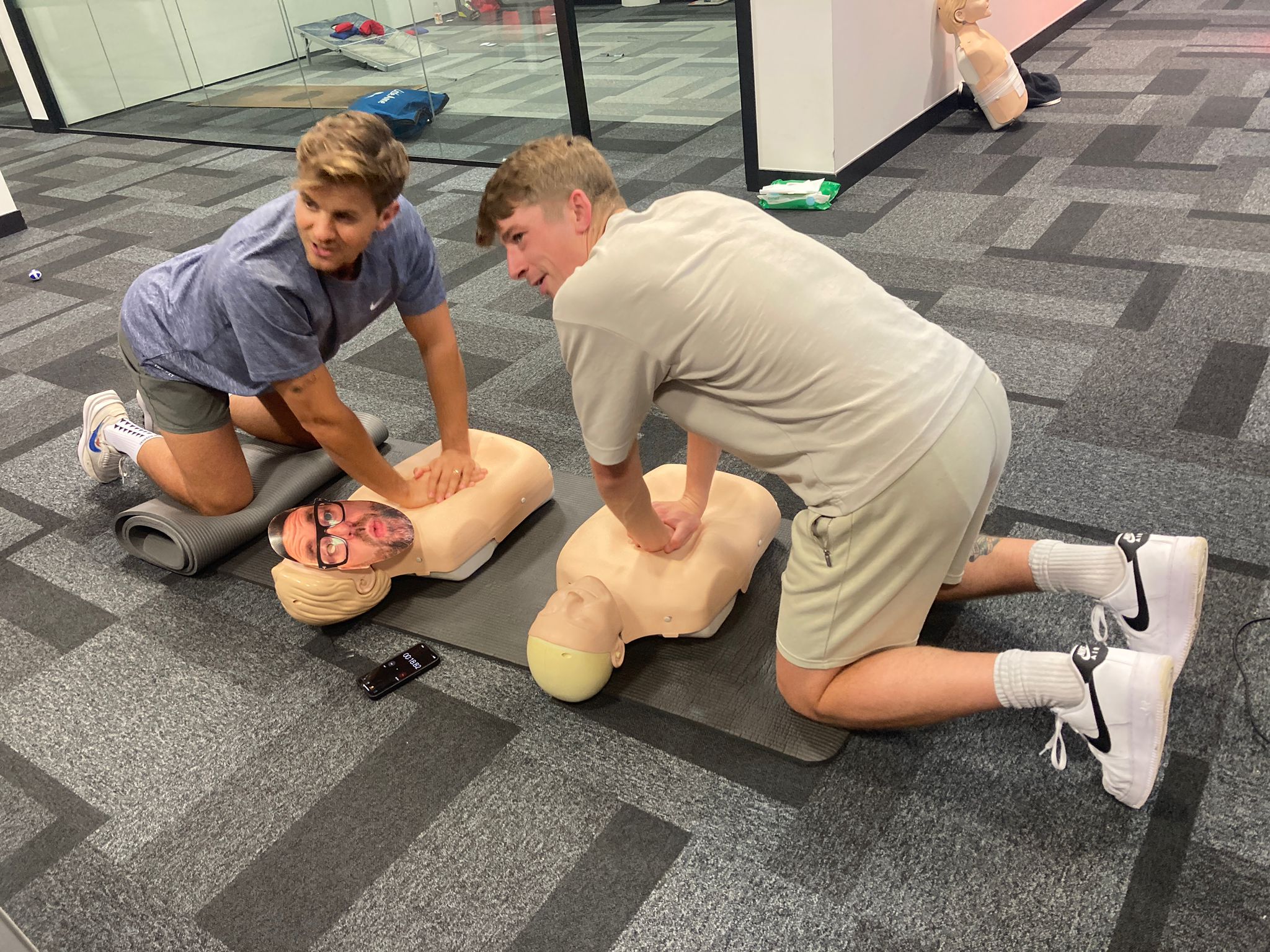 Two men are kneeling on an office floor, leaning forward with their hand of the chest of CPR mannikins.