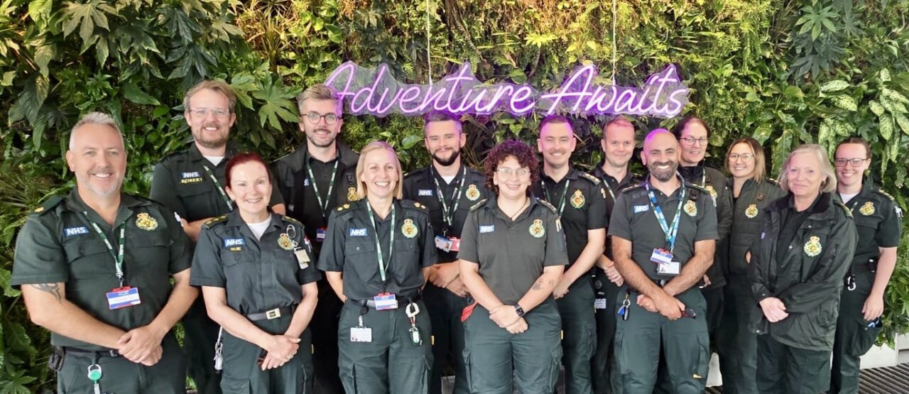 A group of male and female ambulance staff in green uniforms stand in a group for a publicity photo.