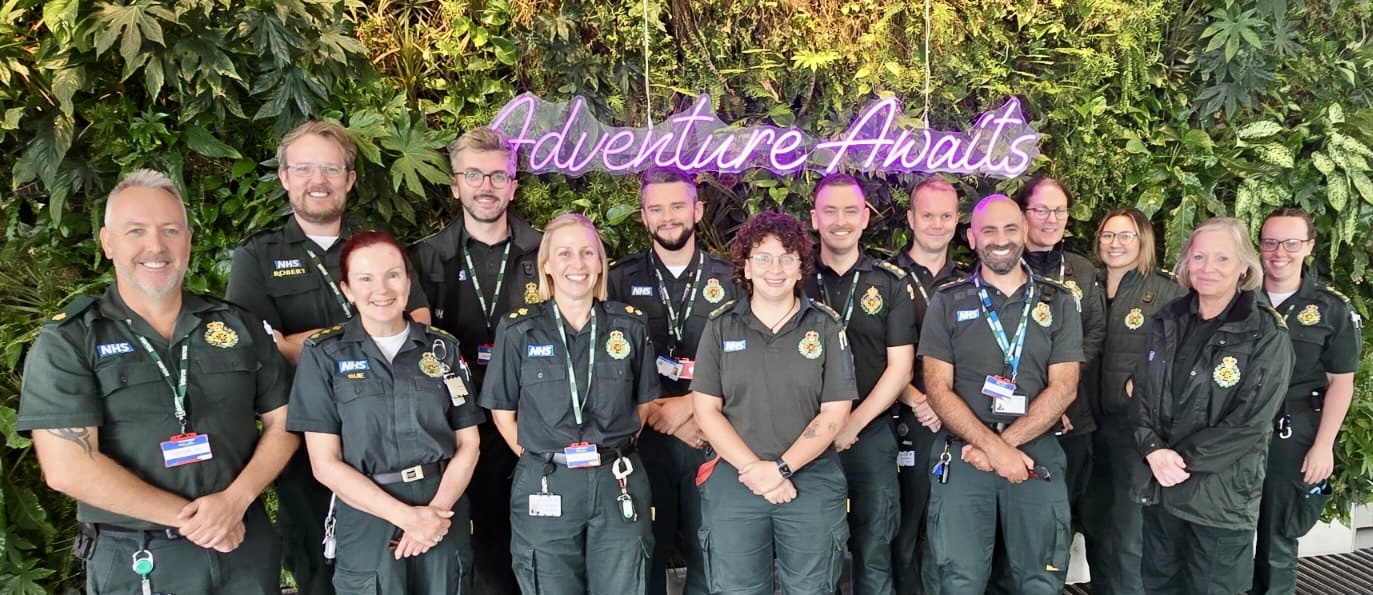 A group of male and female ambulance staff in green uniforms stand in a group for a publicity photo.