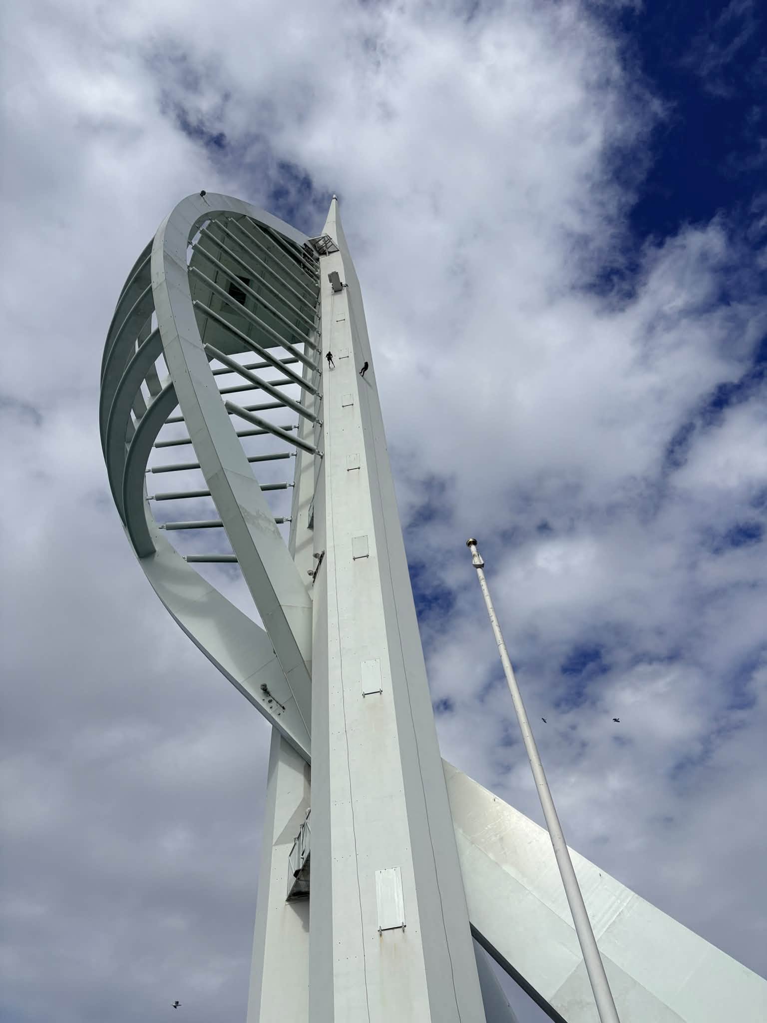 Two people are seen descending The Spinnaker Tower in Portsmouth by abseiling. The photo is taken from the base of the tower looking up.